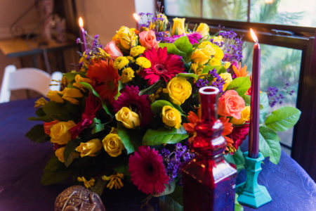 Floral table centerpiece with red, pink and orange Gerbera Daisy's, peach - yellow roses, filler flowers on an indigo tablecloth with purple glass bottle, lit candles in dark turquoise holders.