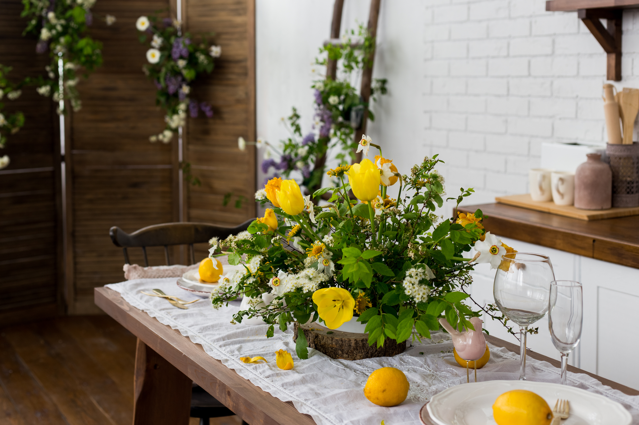 A bouquet of yellow tulips, white flowers with leaves decorates a served wooden table with a white tablecloth. On the table are lemons, plates, glasses. Stylish living room decor in Scandinavian style