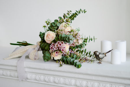 Beautiful flower wedding bouquet of white roses, pink roses, with sprigs of eucalyptus on white wooden table with white candles and metal bird decoration lying on fireplace